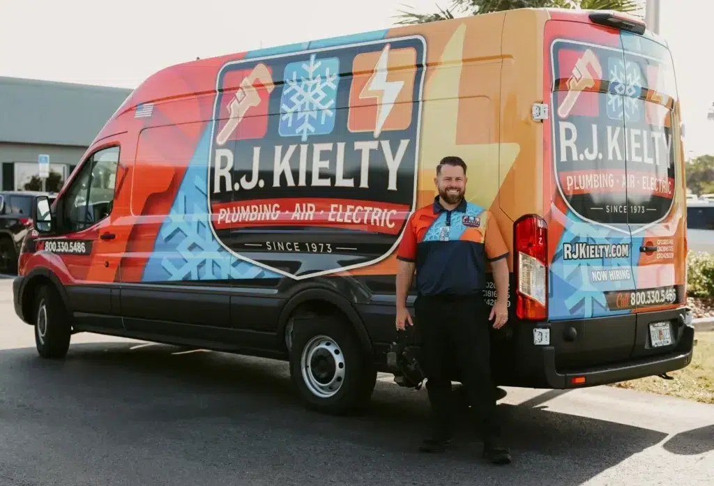 A smiling RJ Kielty technician standing proudly next to a fully branded service van equipped for plumbing, air conditioning and electrical repairs